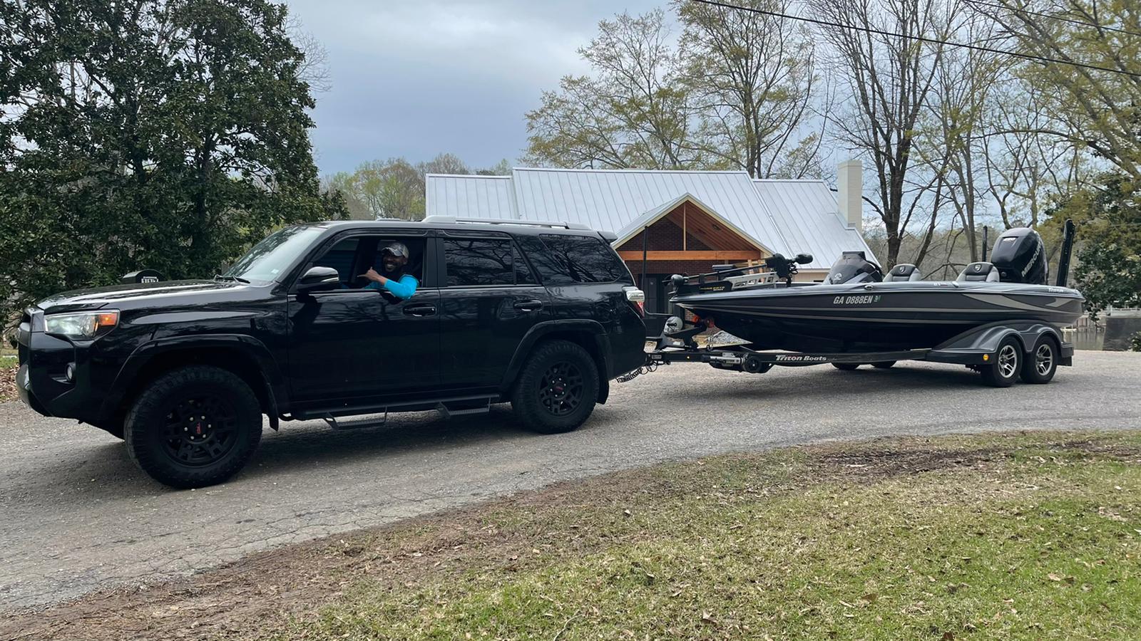 Black SUV towing a boat on a gravel driveway. A person is driving. House and trees in the background.