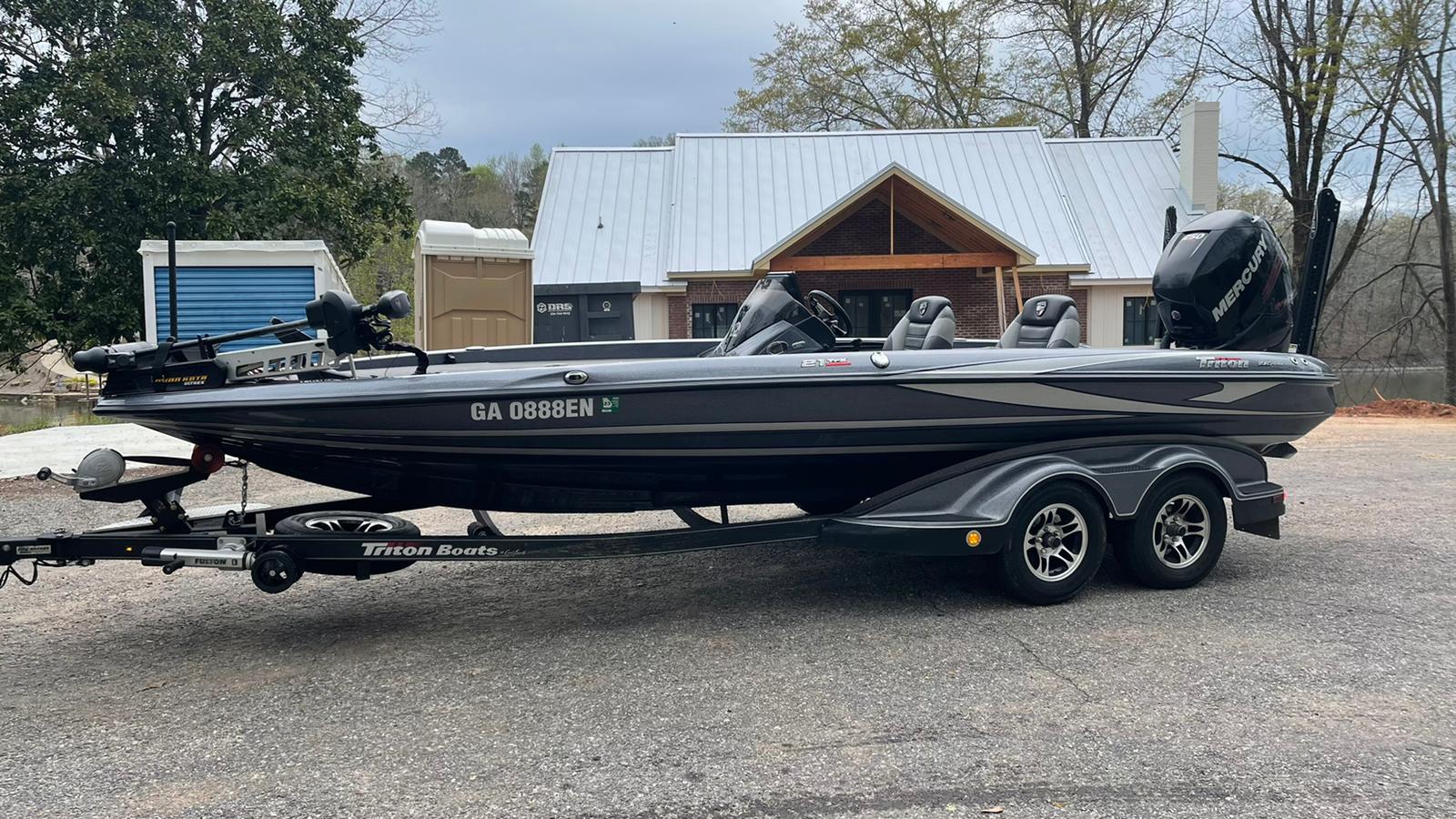 Dark grey bass fishing boat on a trailer; set in front of a building with a tin roof.