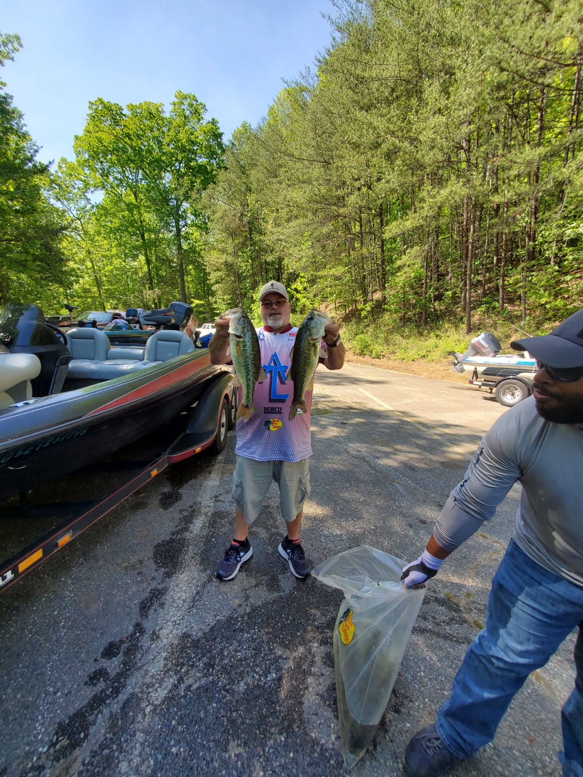 Man holding two bass fish, with a person holding a bag. Boats and trees in the background.