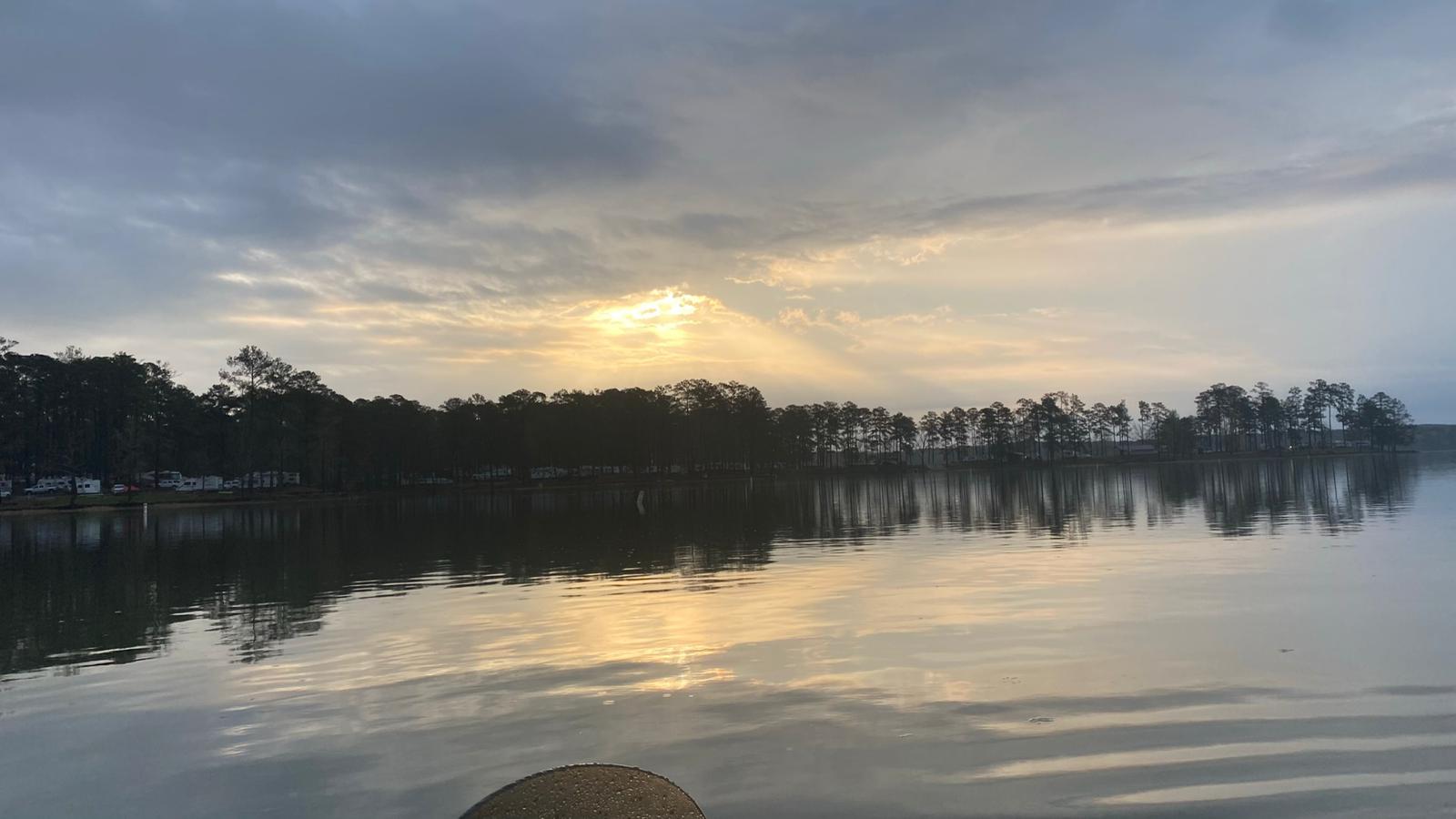 Overcast sky with sunlight breaking through, reflecting on calm water and shoreline trees.