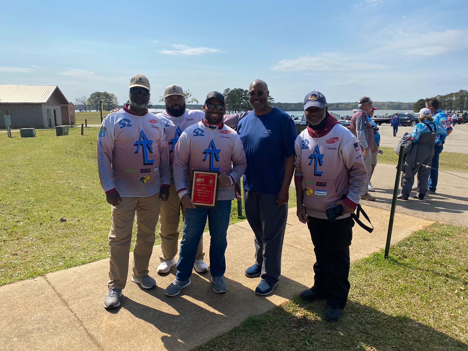 Five men in matching shirts hold a plaque outdoors.