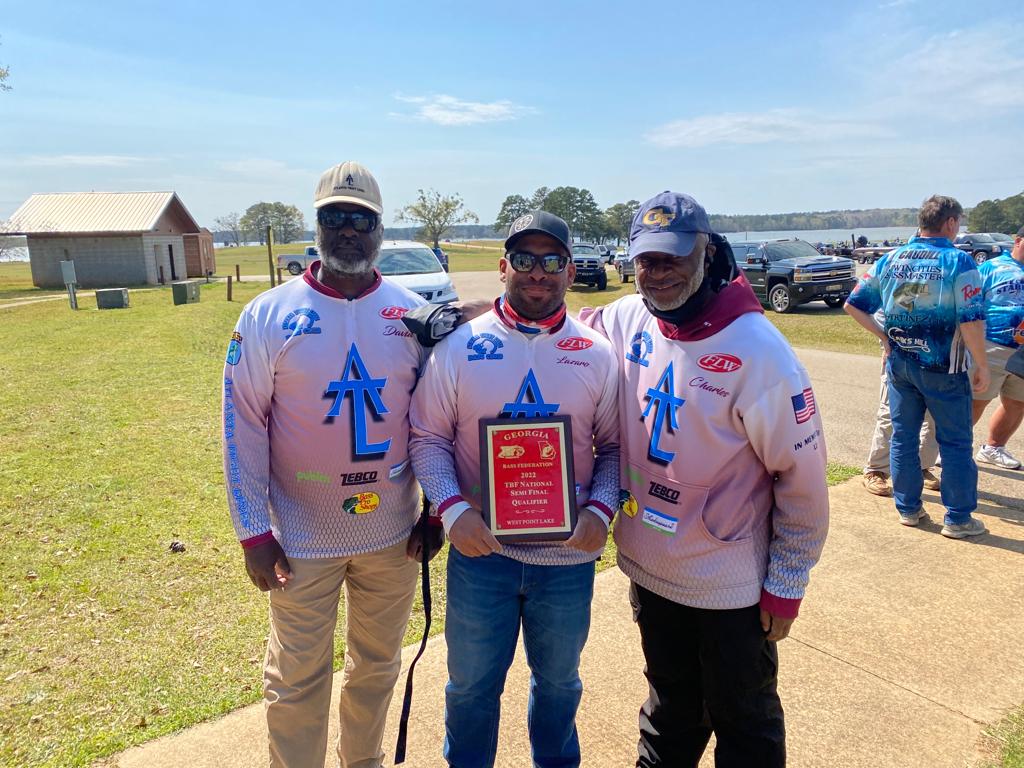 Three men holding a trophy outside, wearing fishing team shirts, smiling.