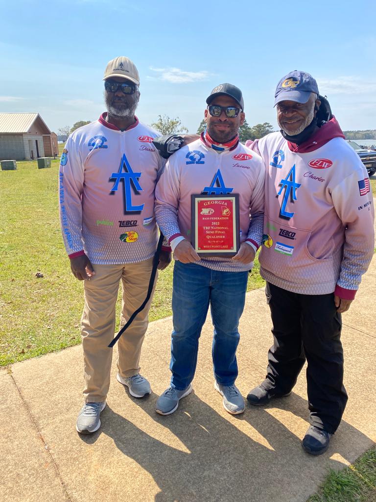 Three men in matching pink jerseys stand outside, holding a trophy.