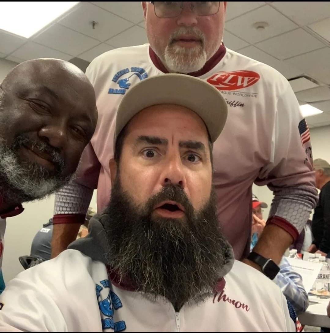 Three men in a room, one taking a selfie with a surprised expression, other two smiling; a white shirt with