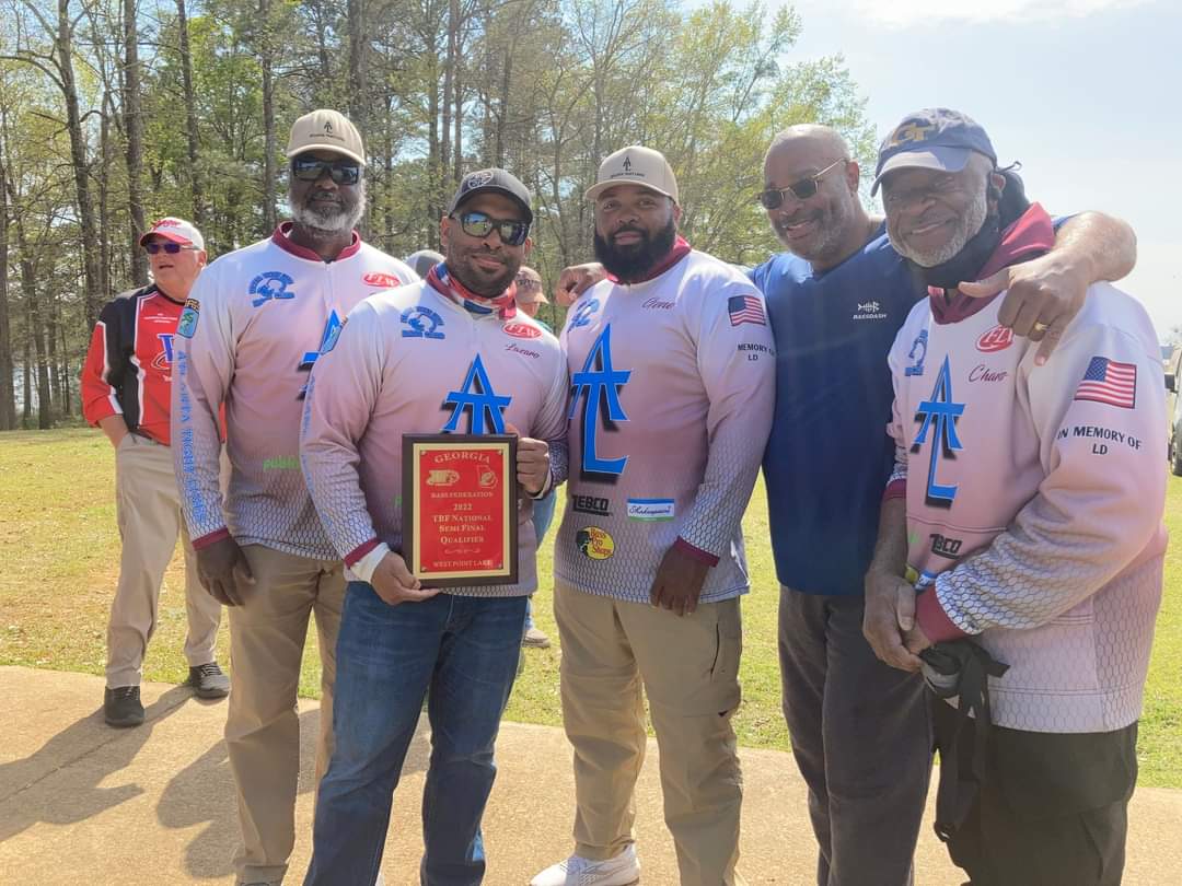 Group of men in matching shirts holding a trophy outdoors. Some have arms around each other, smiling.