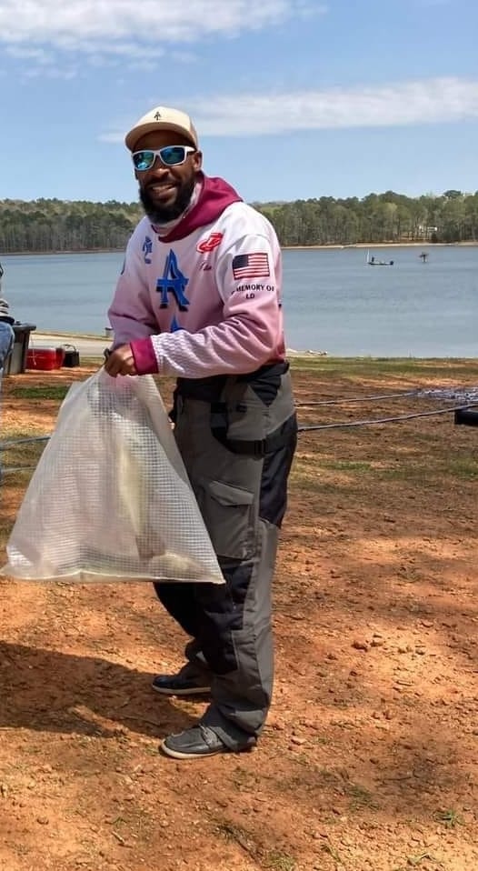 Man in pink hoodie holds a clear bag on a lakeside shore, smiling.