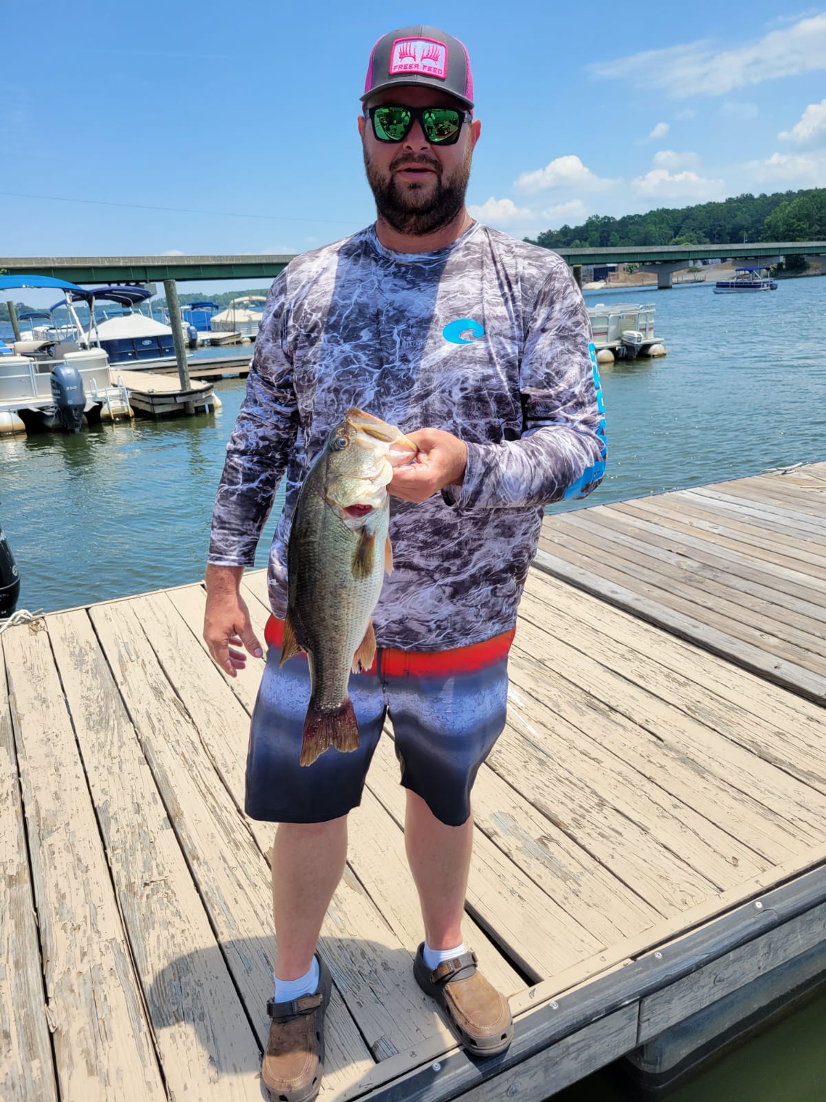 Man holding a fish on a dock, lake in background. He wears a hat, sunglasses, long-sleeve shirt, and shorts.