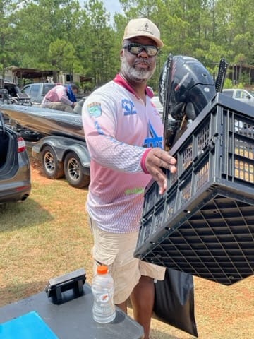 Man holding a black crate near a boat. He wears sunglasses, a hat, and a long-sleeved shirt. Outdoors.
