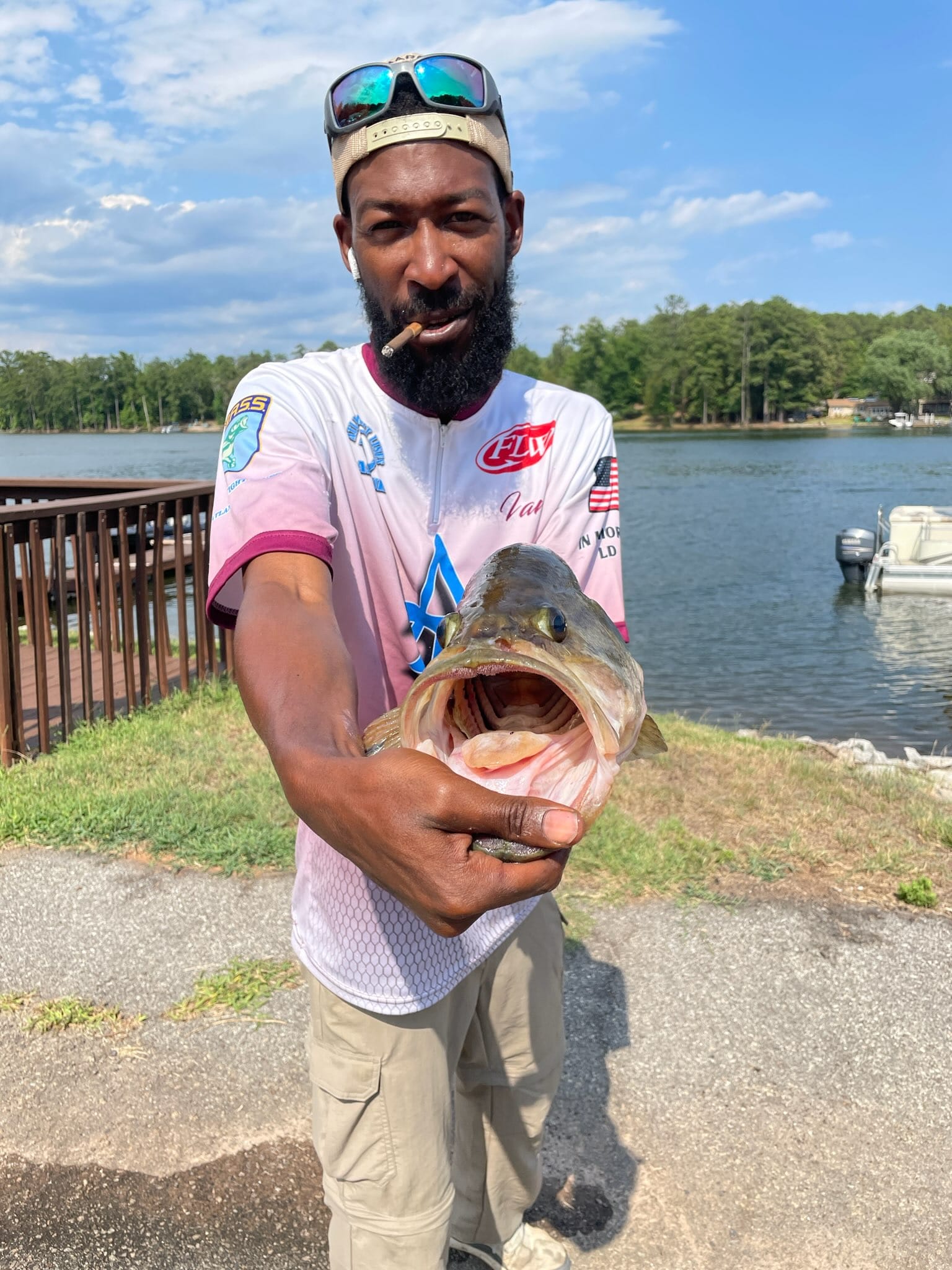 A man holding a large fish with its mouth open wide near a lake. He wears a hat, sunglasses, and a jersey.