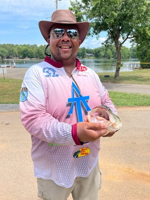 Man holding a fish, wearing a hat and sunglasses, smiles near a lake.