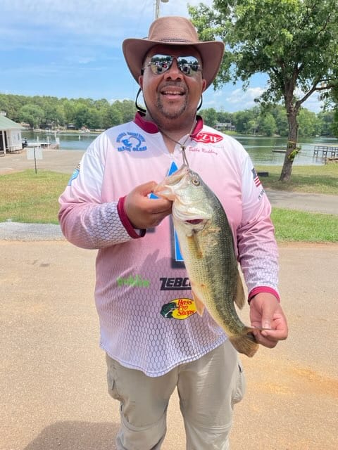 Man in hat holding a largemouth bass. He is smiling, standing outside near a lake.