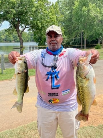 Man holding up two large fish, lake background. He wears a pink shirt and a hat, smiling.