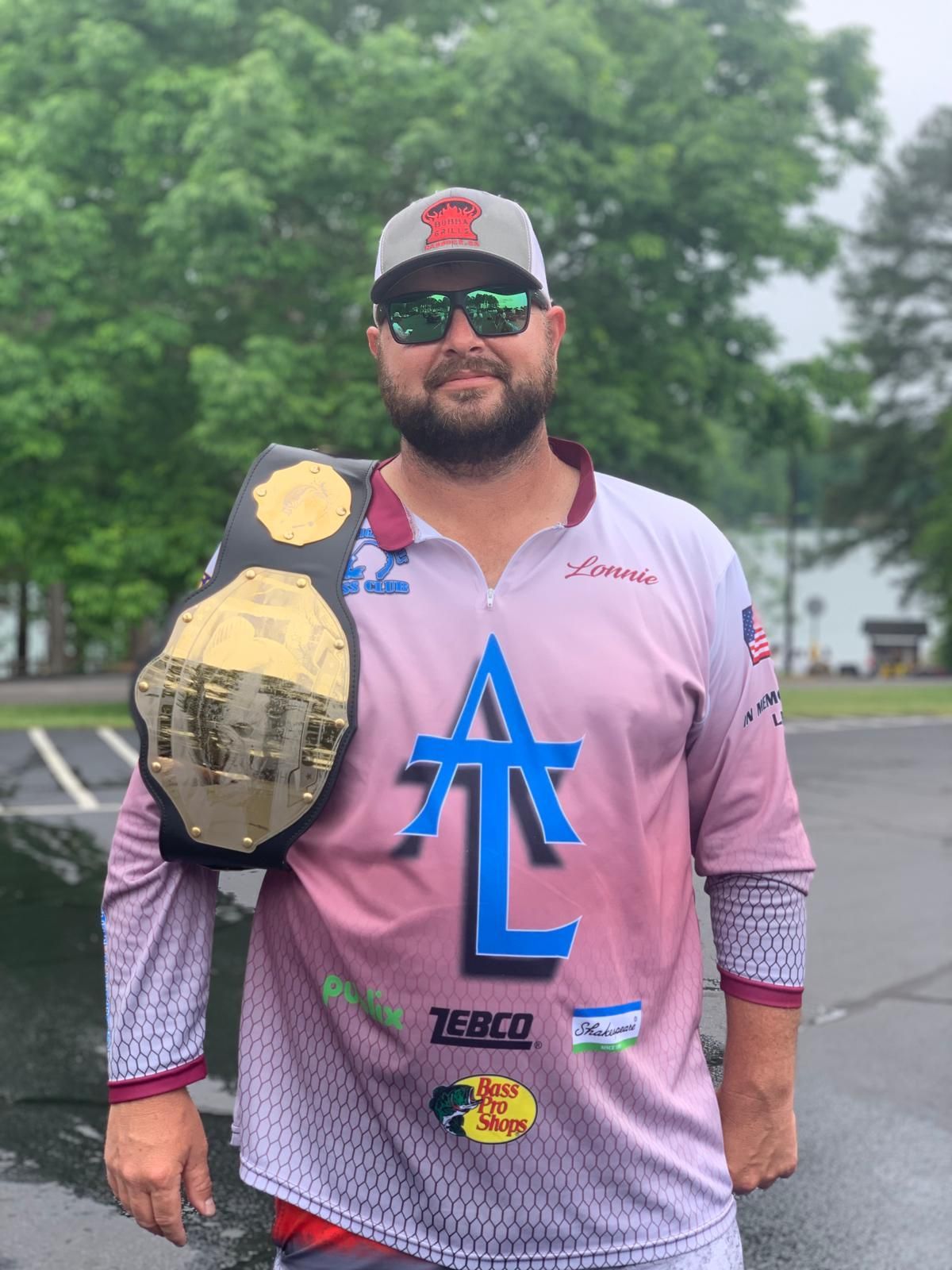 Man in fishing shirt holding a championship belt. Gray hat, sunglasses, and outdoor setting.