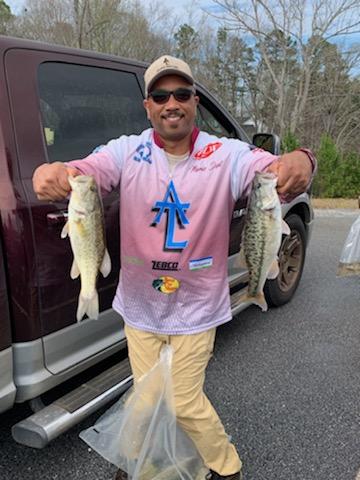 Man holding two bass fish, smiling, near a truck. He wears a fishing shirt, khaki pants, and a cap.