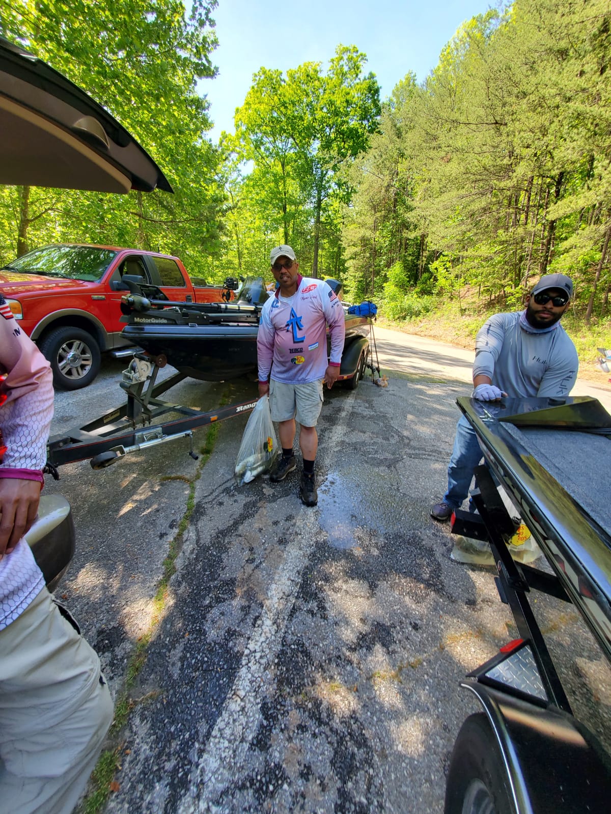 People at a boat ramp. Man in pink shirt stands next to a boat, another person cleans. Red truck in background.