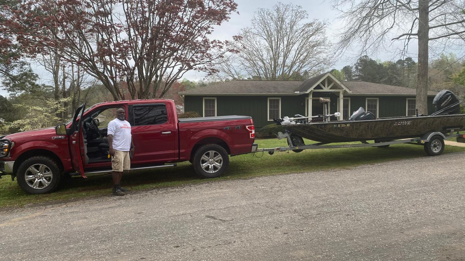 Man beside red truck towing boat on a trailer in front of a house.