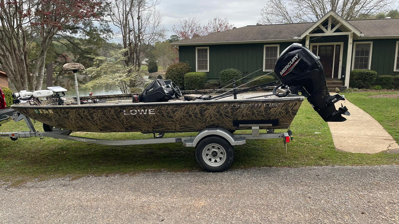Camouflage boat on a trailer in front of a green house, with a black outboard motor.