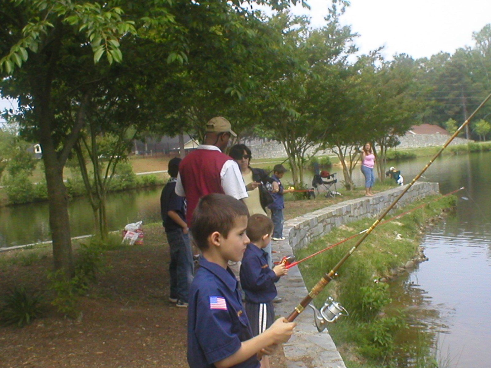 Children fishing at a pond, trees in background. Boy Scouts in uniform, adults present.
