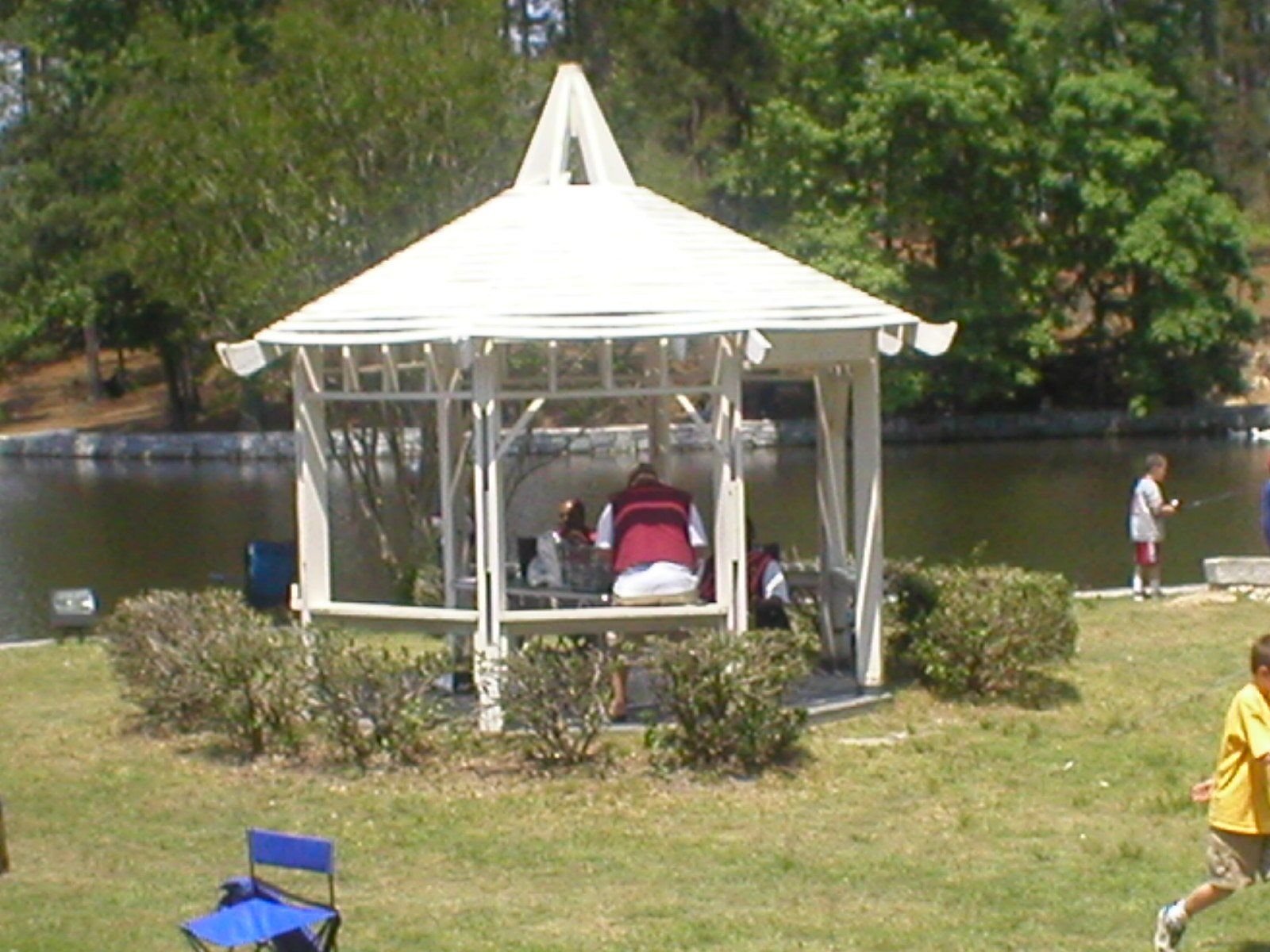White gazebo with people inside, near a lake; children fishing nearby.