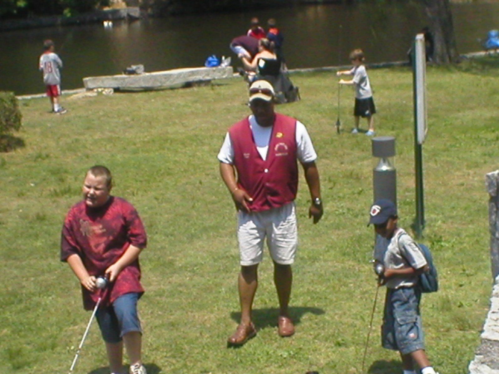 People fishing at a lake; an instructor with children. Sunny day, green grass.
