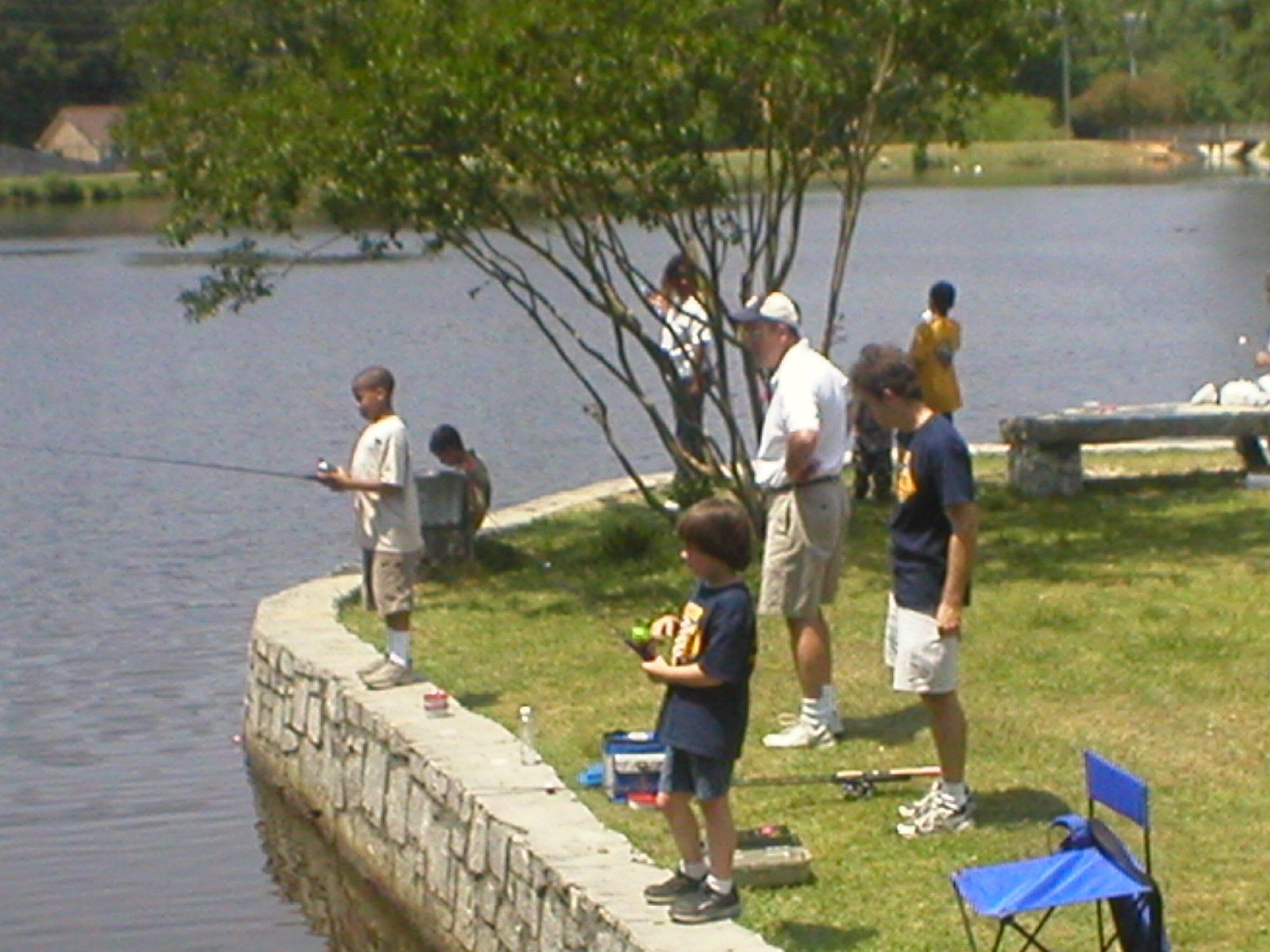 People fishing by a lake on a sunny day. Several children and adults near the water.