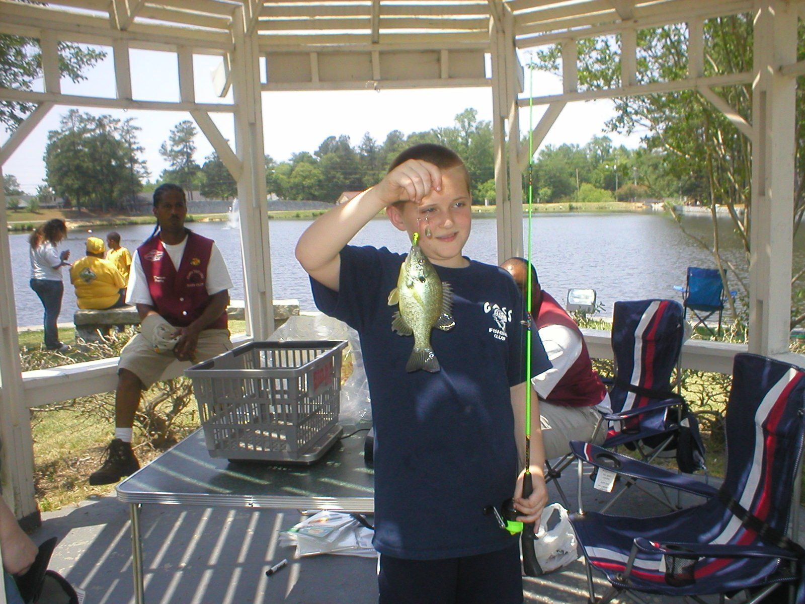 Boy in gazebo holds up small fish, smiling. Lake and other people in background.