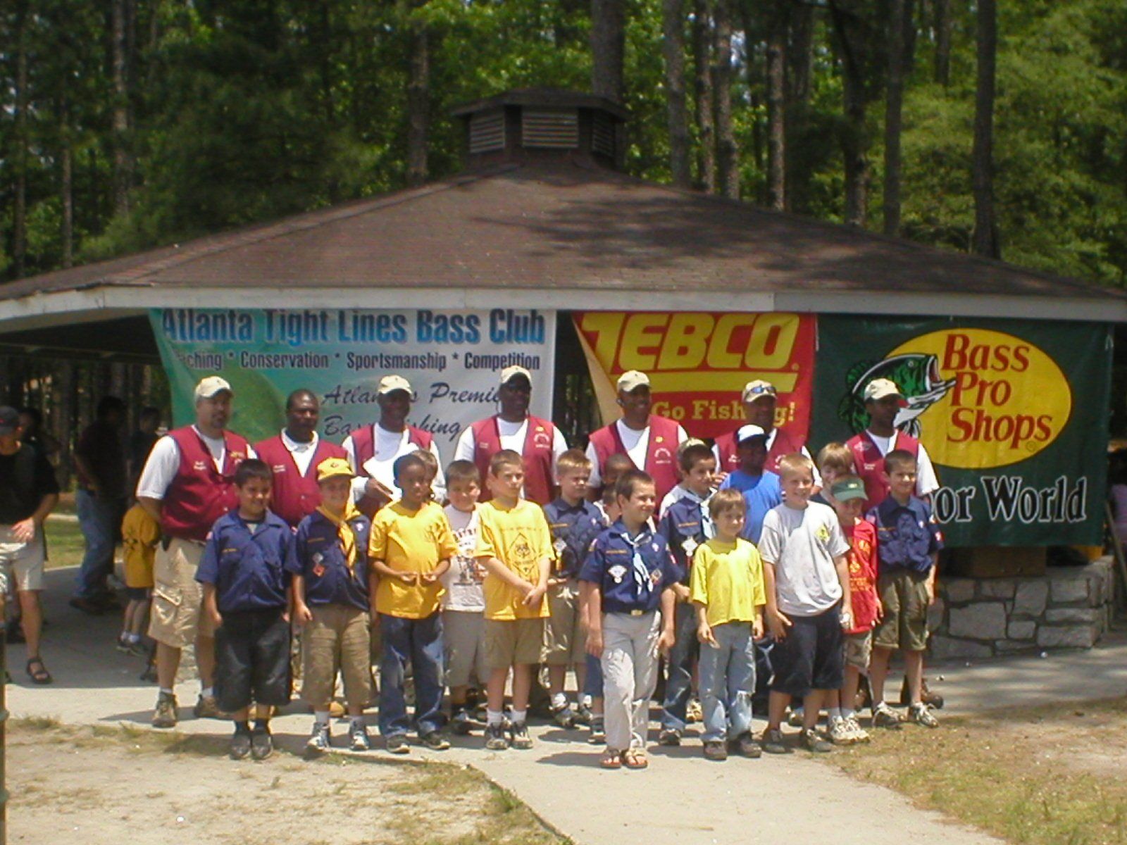 Group of children and adults in vests pose in front of a gazebo with banners for fishing.