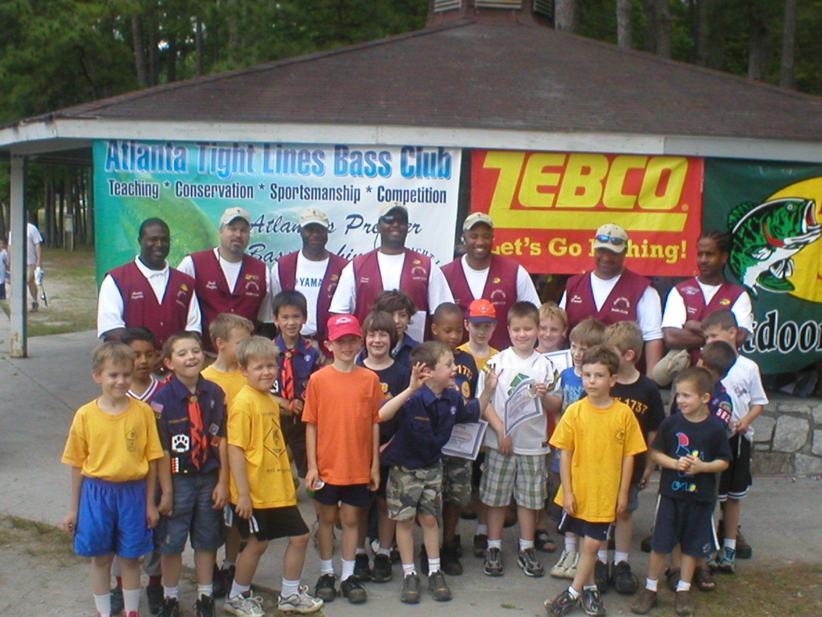Group photo of adults and children at a fishing event, standing in front of a gazebo and banner.