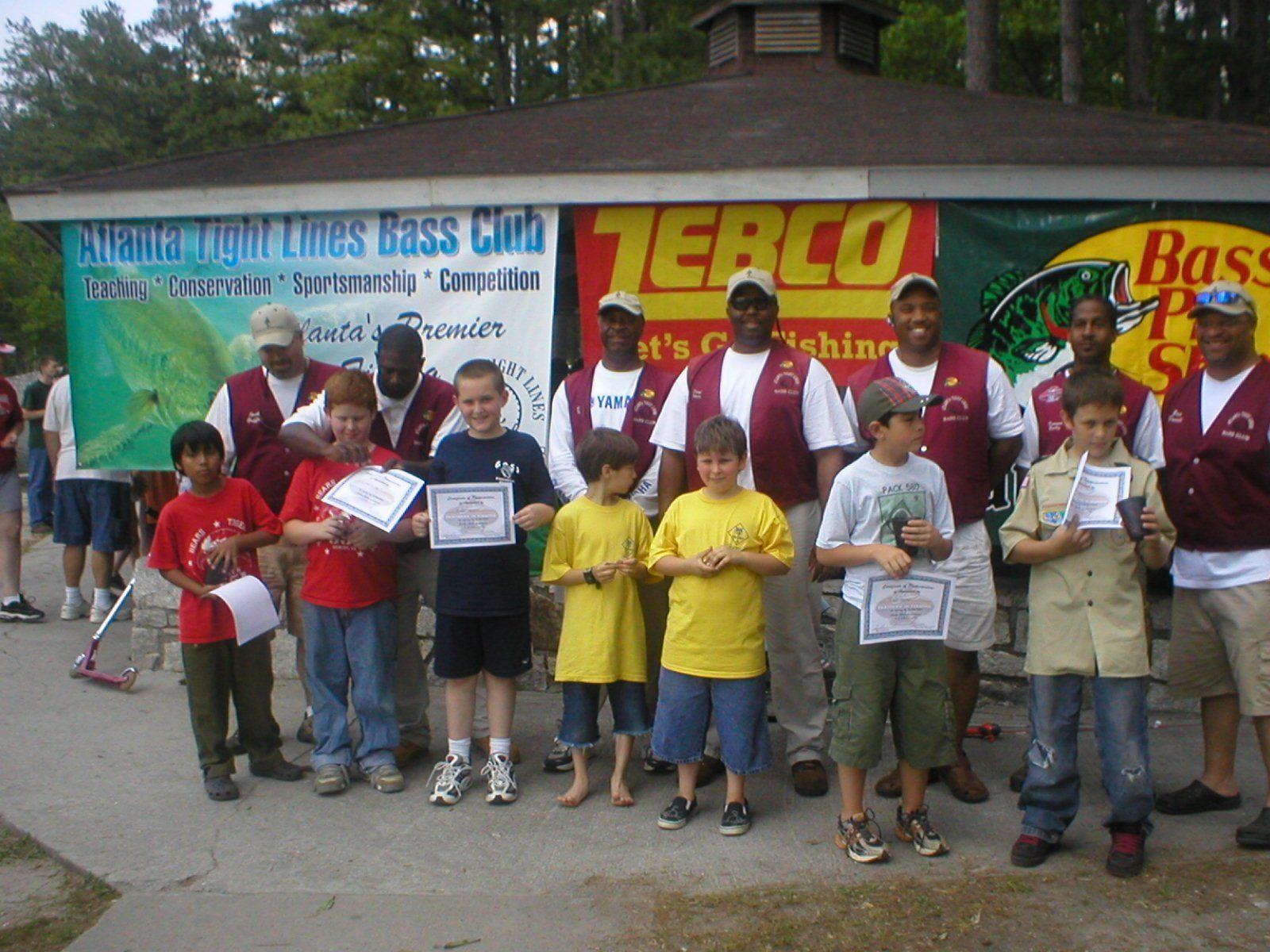 Children and adults at a fishing competition posing with trophies and certificates near a building with banners.