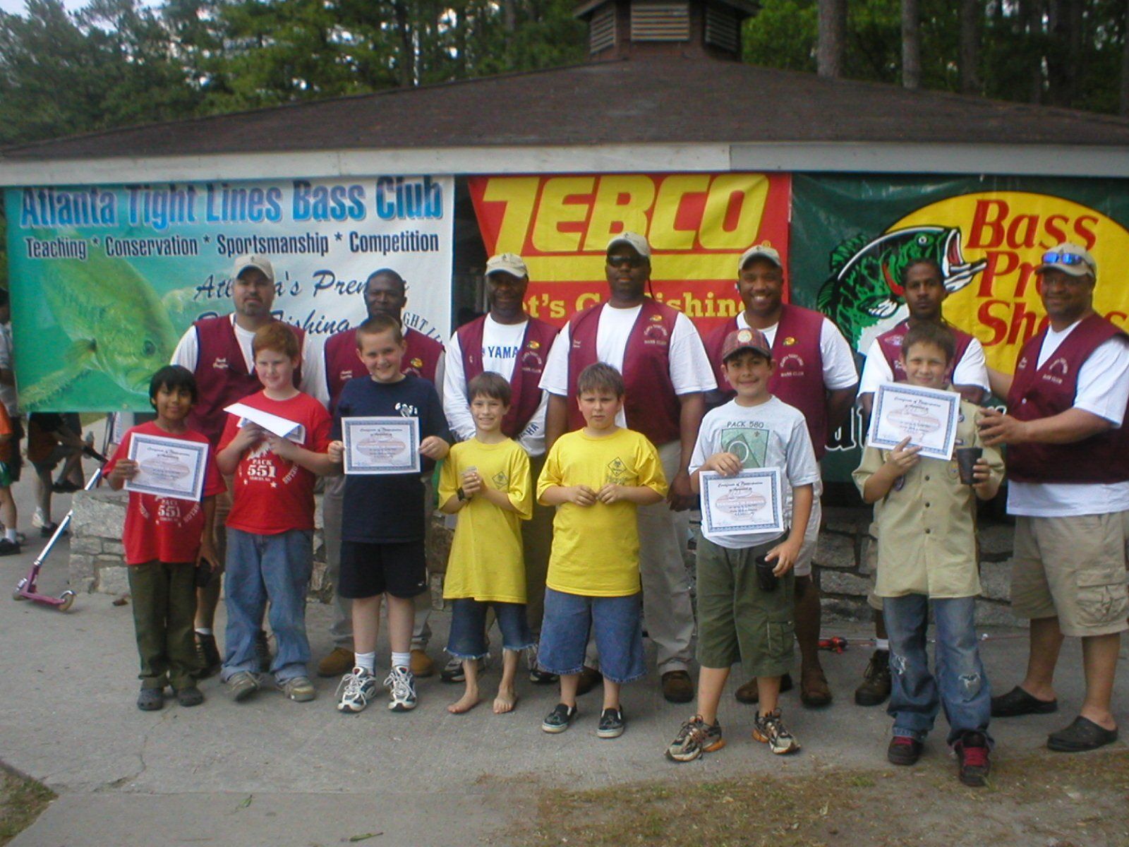 Kids and adults holding certificates pose in front of sponsor banners after fishing event.