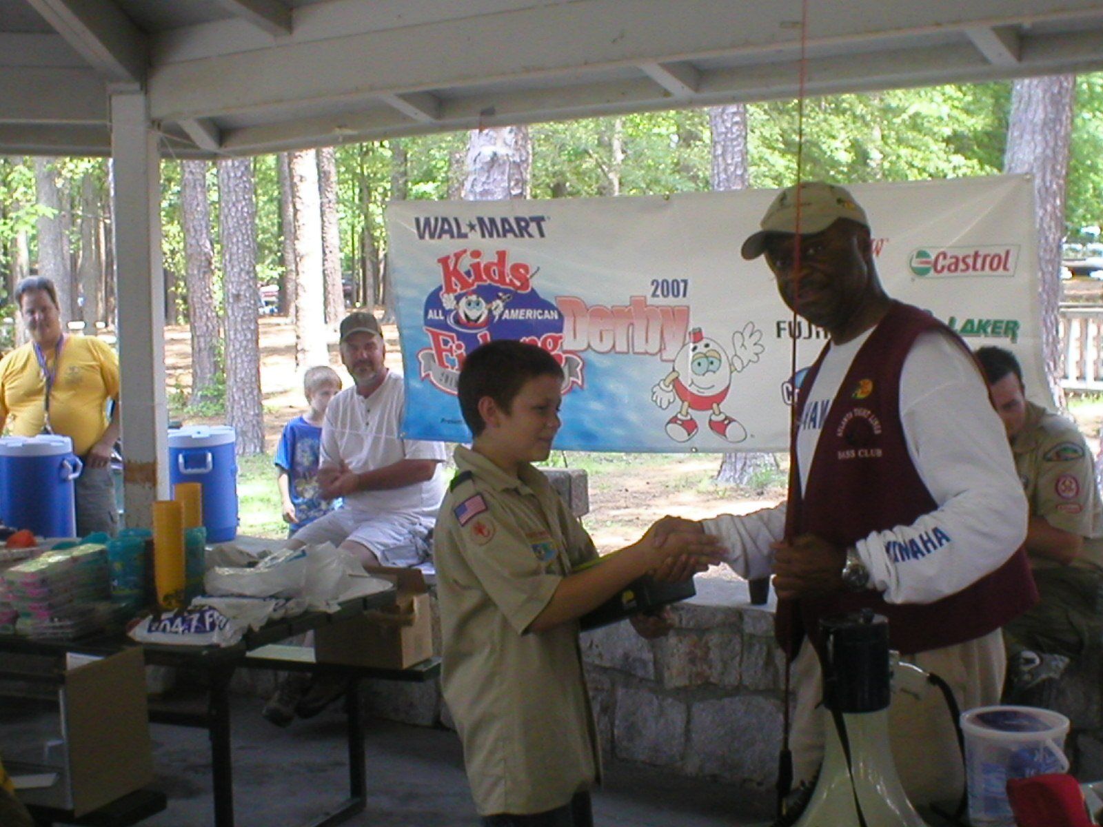 Boy Scout receiving award from man at a Walmart Kids Fishing Derby event, outdoors under a pavilion.