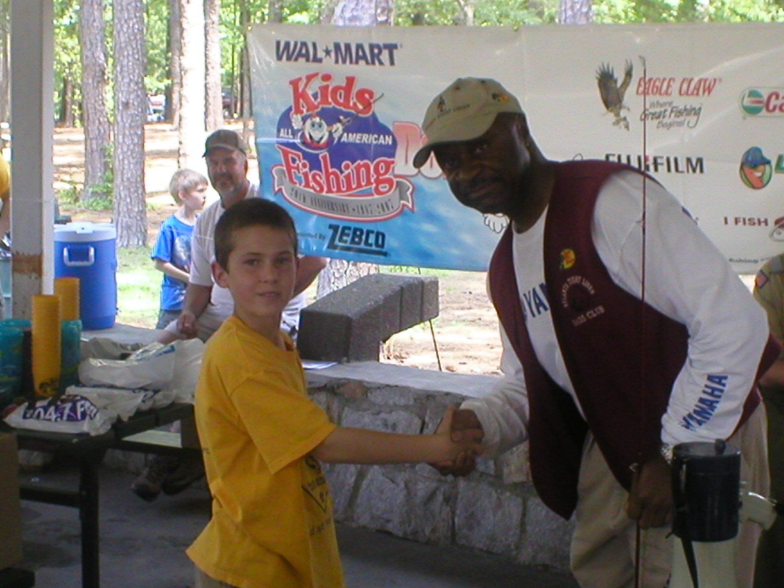 Boy in yellow shirt shakes hands with man at a kids' fishing event sponsored by Walmart.