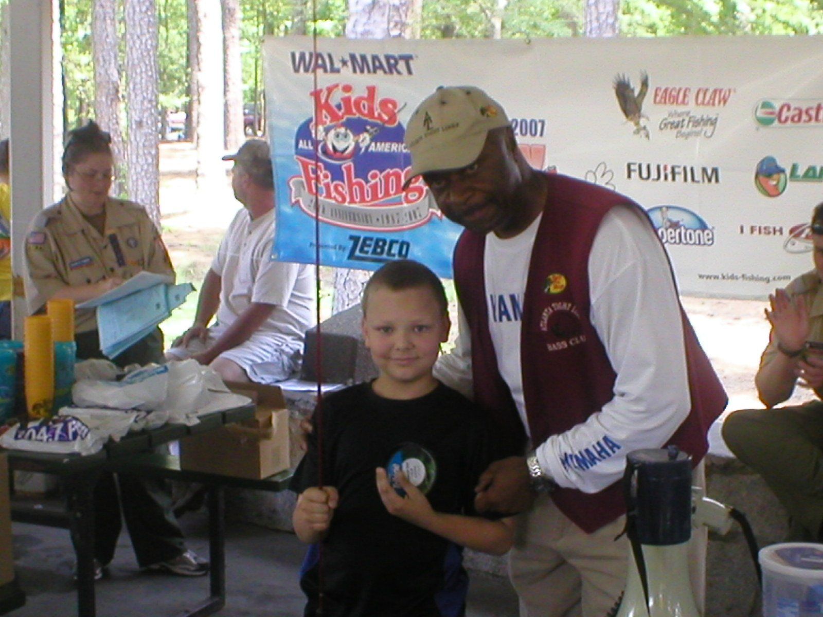 Boy and man at a fishing event. The boy holds a prize, smiles. The man smiles too. Outdoor setting, daytime.