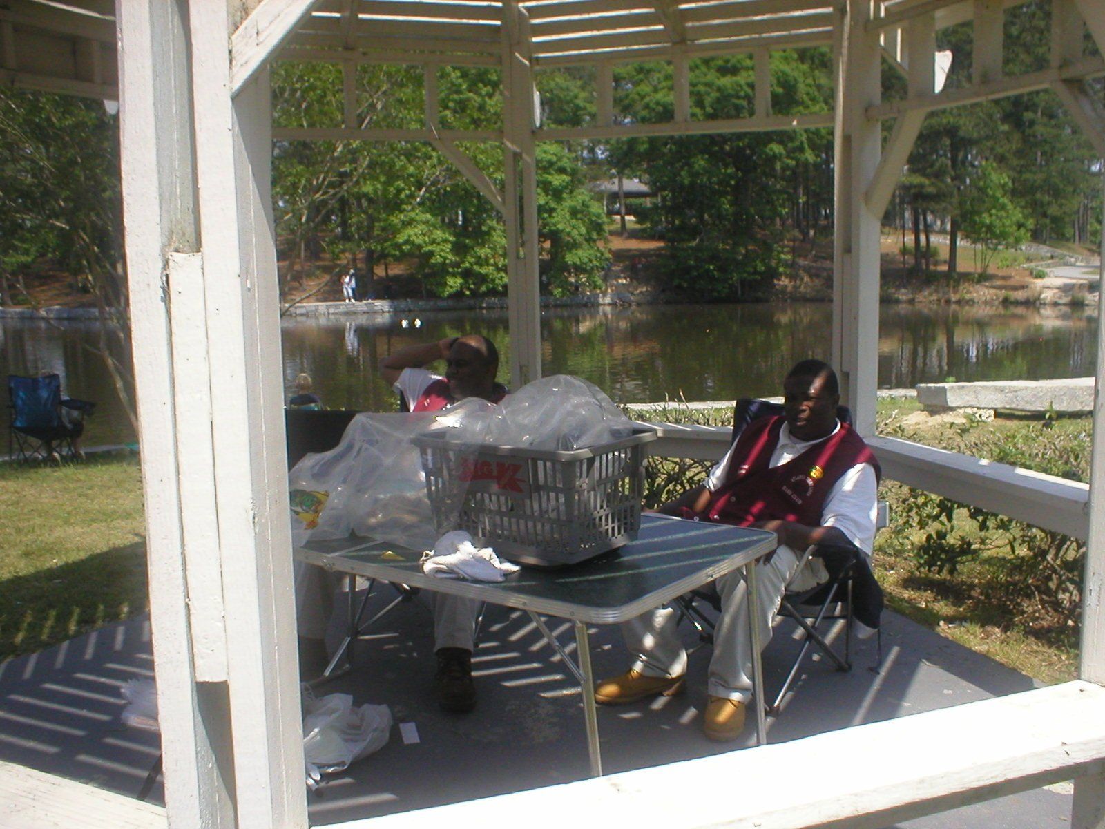 Two people seated at a table in a gazebo by a lake; one is wearing a red vest.