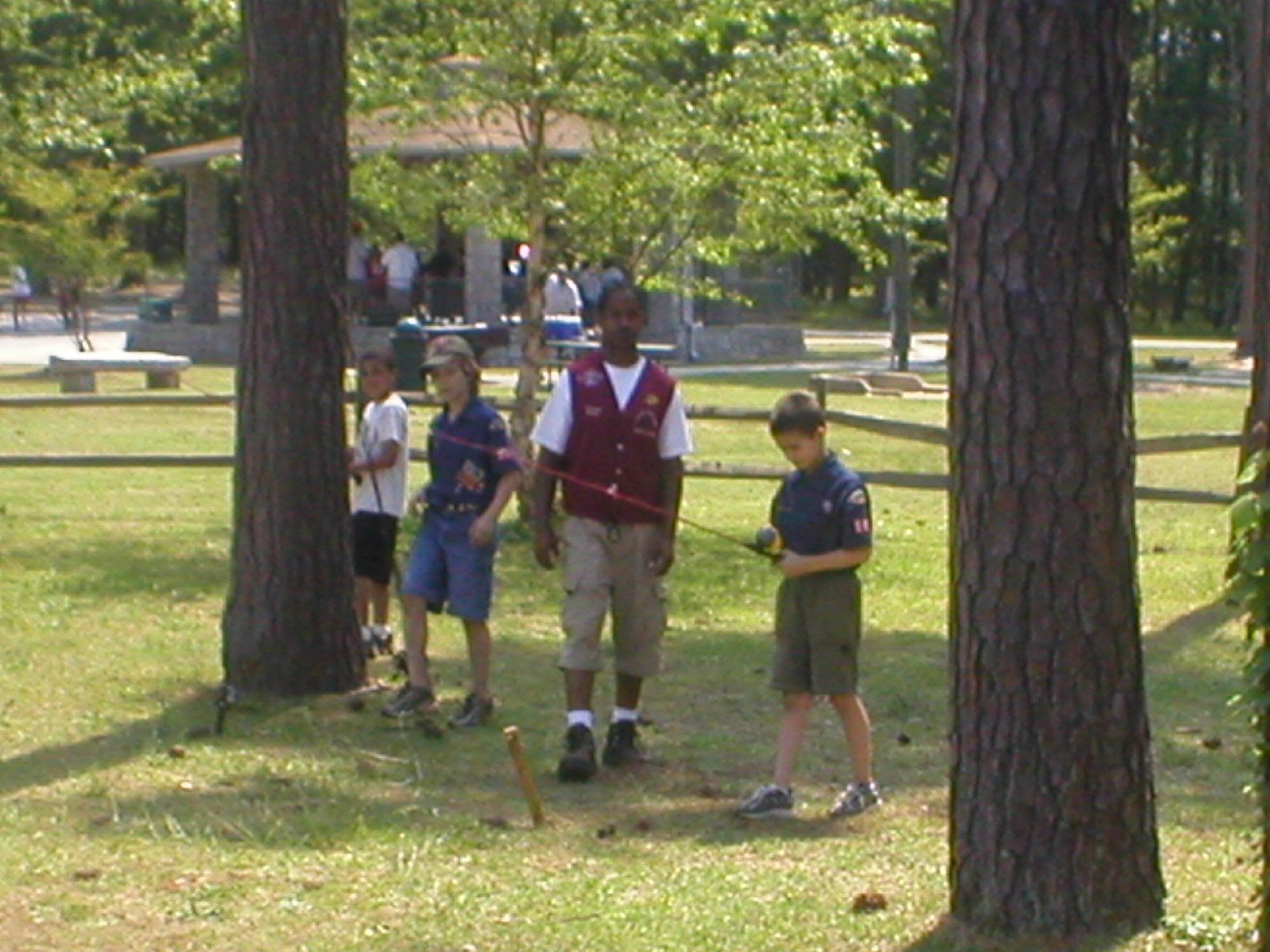 Boy Scouts in a park, walking near trees. A scout leader in a vest guides them; picnic shelter visible.