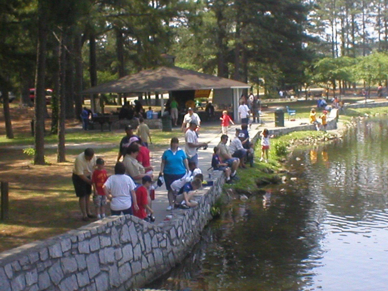 People fishing and gathering near a lake at a park, including a stone bridge.