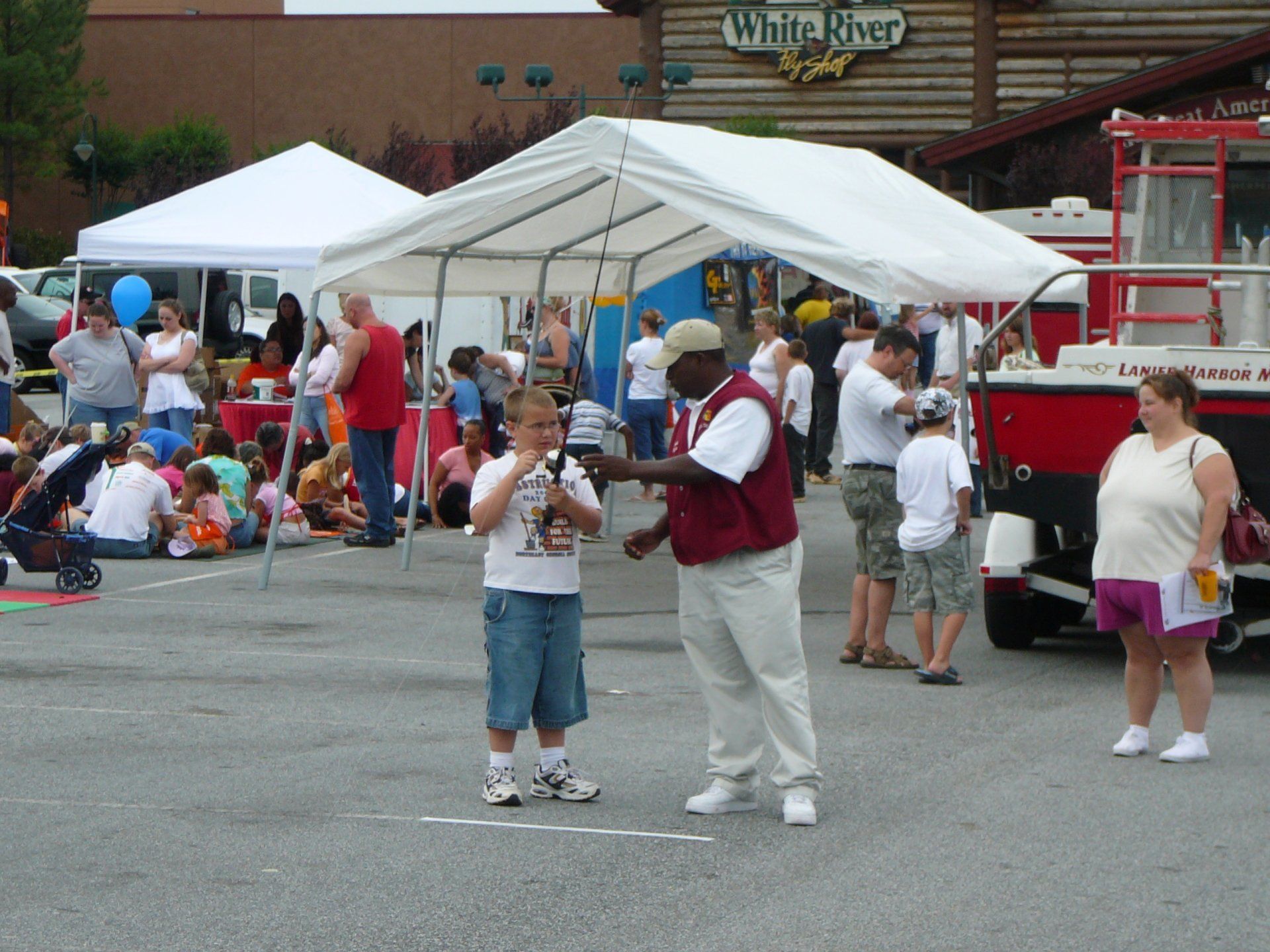 Boy receives trophy from man at outdoor event near a white tent, people, and fire truck.
