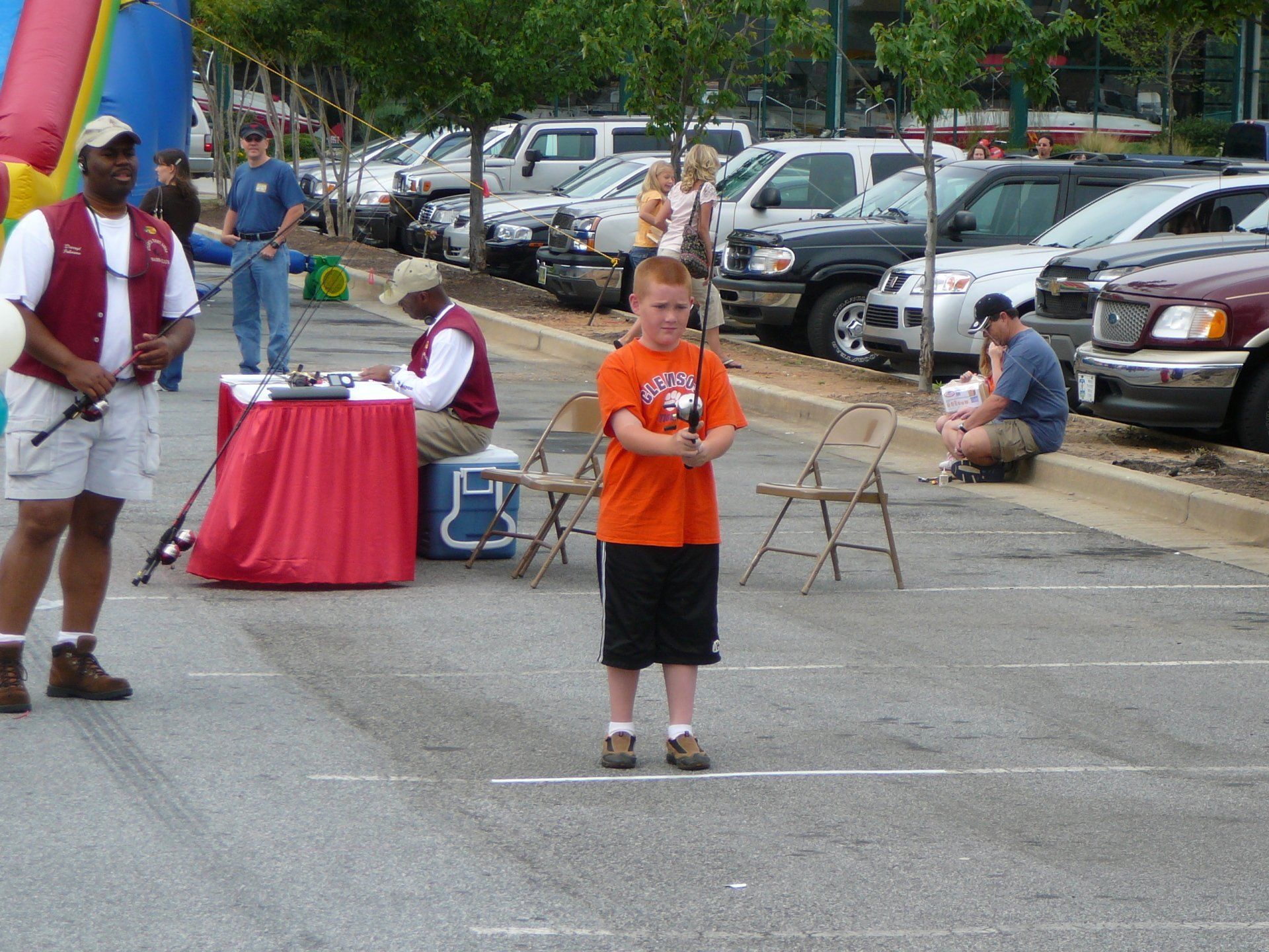 Boy in orange shirt holding fishing rod at outdoor event; parking lot setting.
