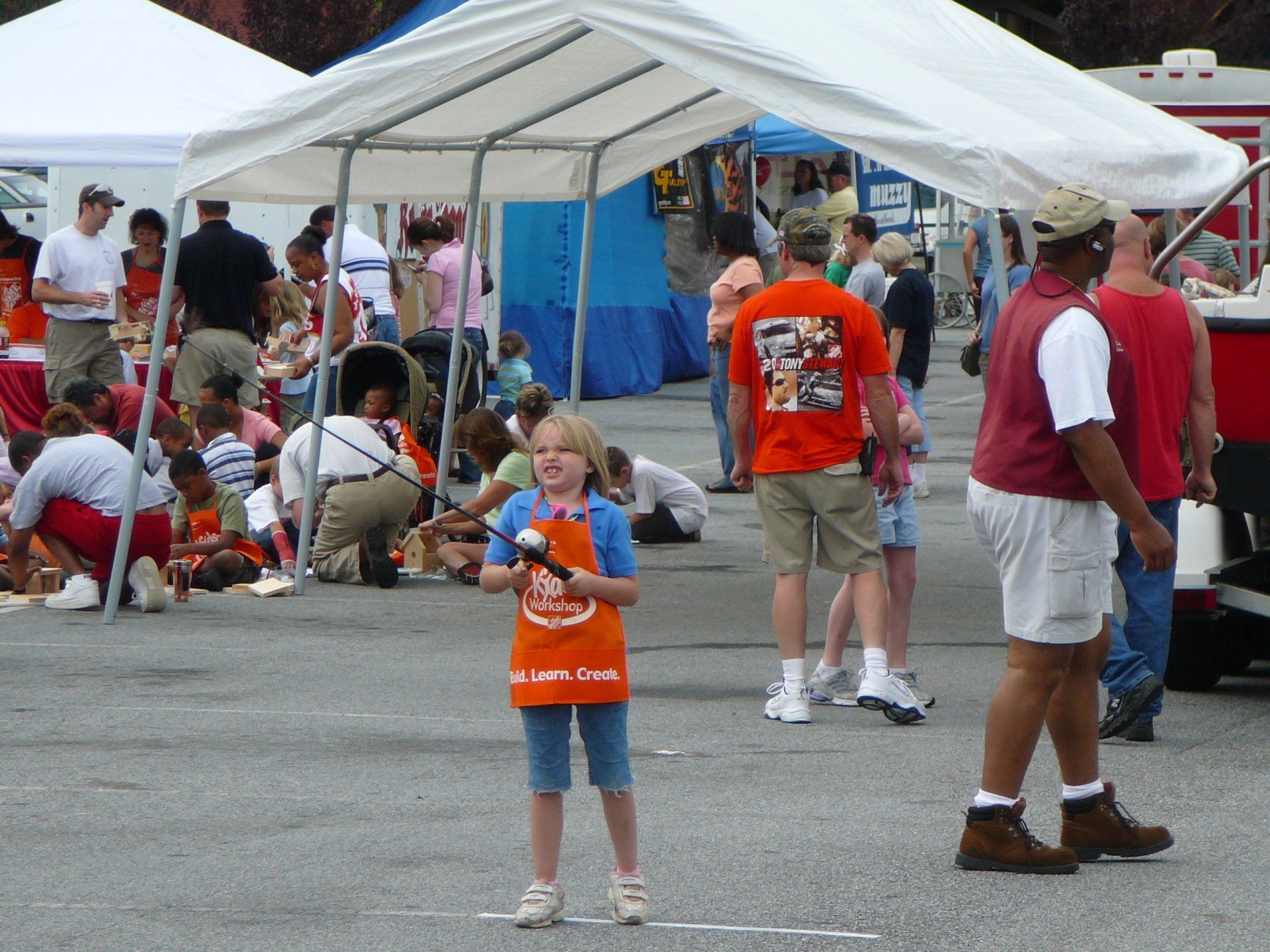 Girl with tool in apron at outdoor event; crowd and tents in the background.