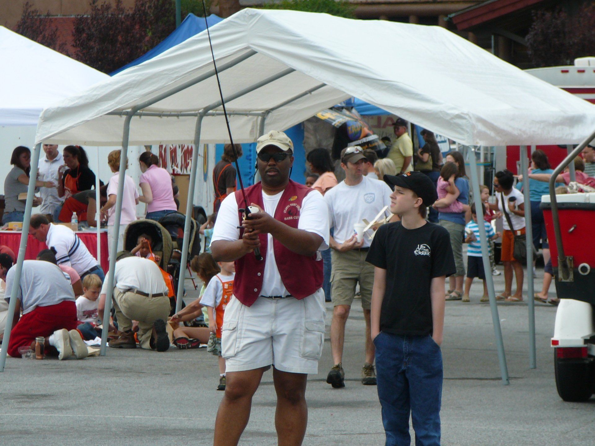 Man in vest and shorts holding a fishing rod, teaching a boy outdoors under a tent.