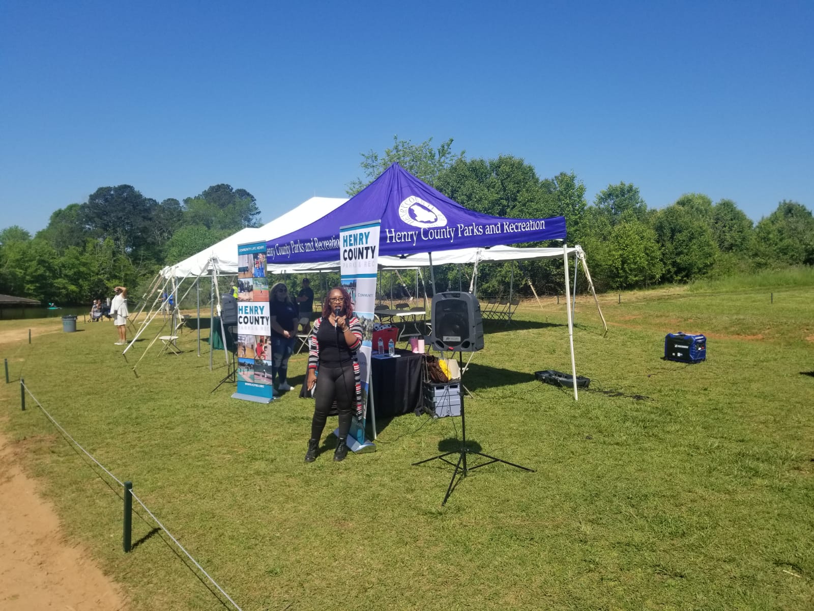 Woman speaking at a booth under a tent with blue branding in a grassy park on a sunny day.