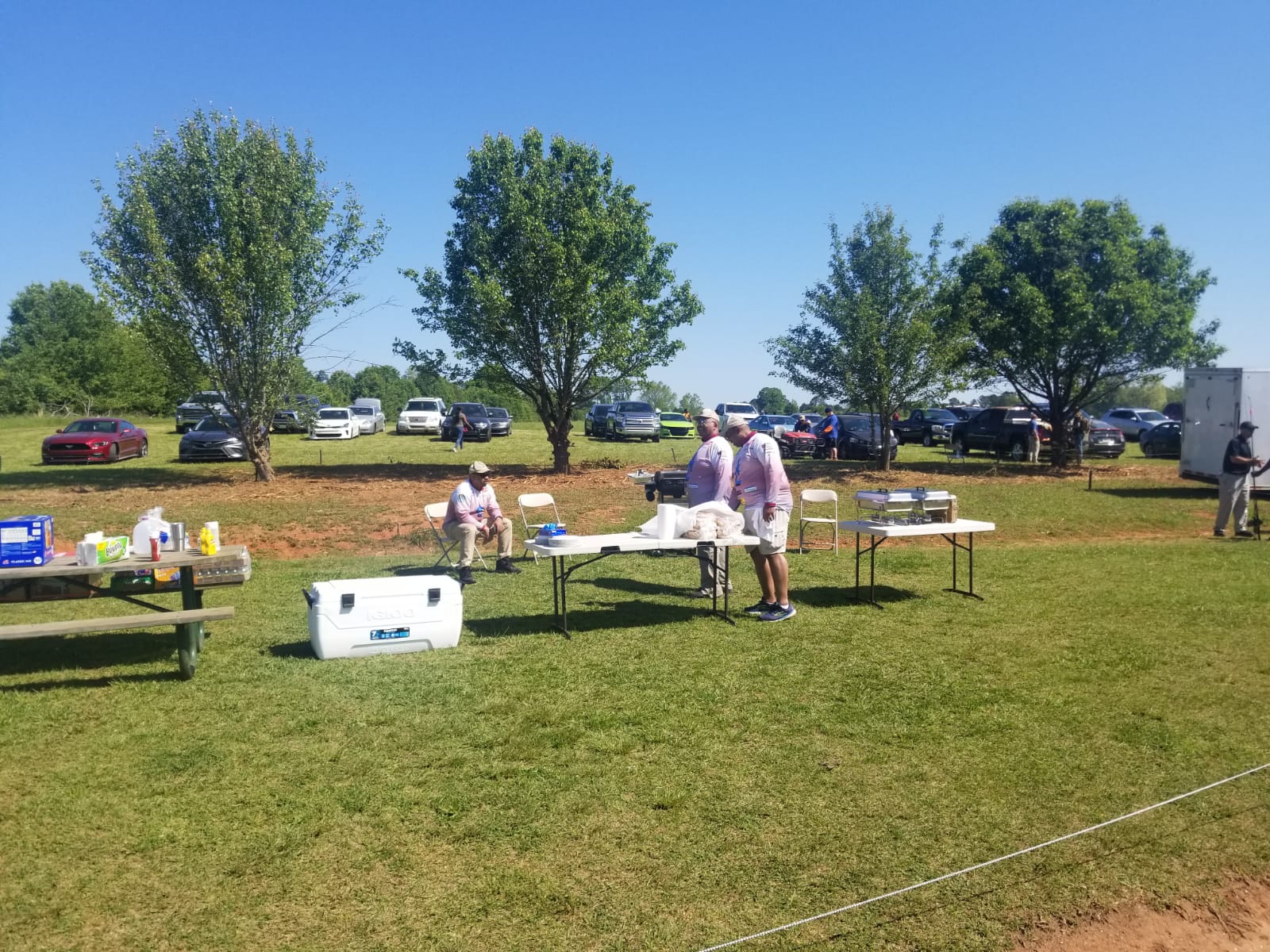 People at a picnic with tables, coolers, and food outdoors. Cars are parked in a grassy field with trees.