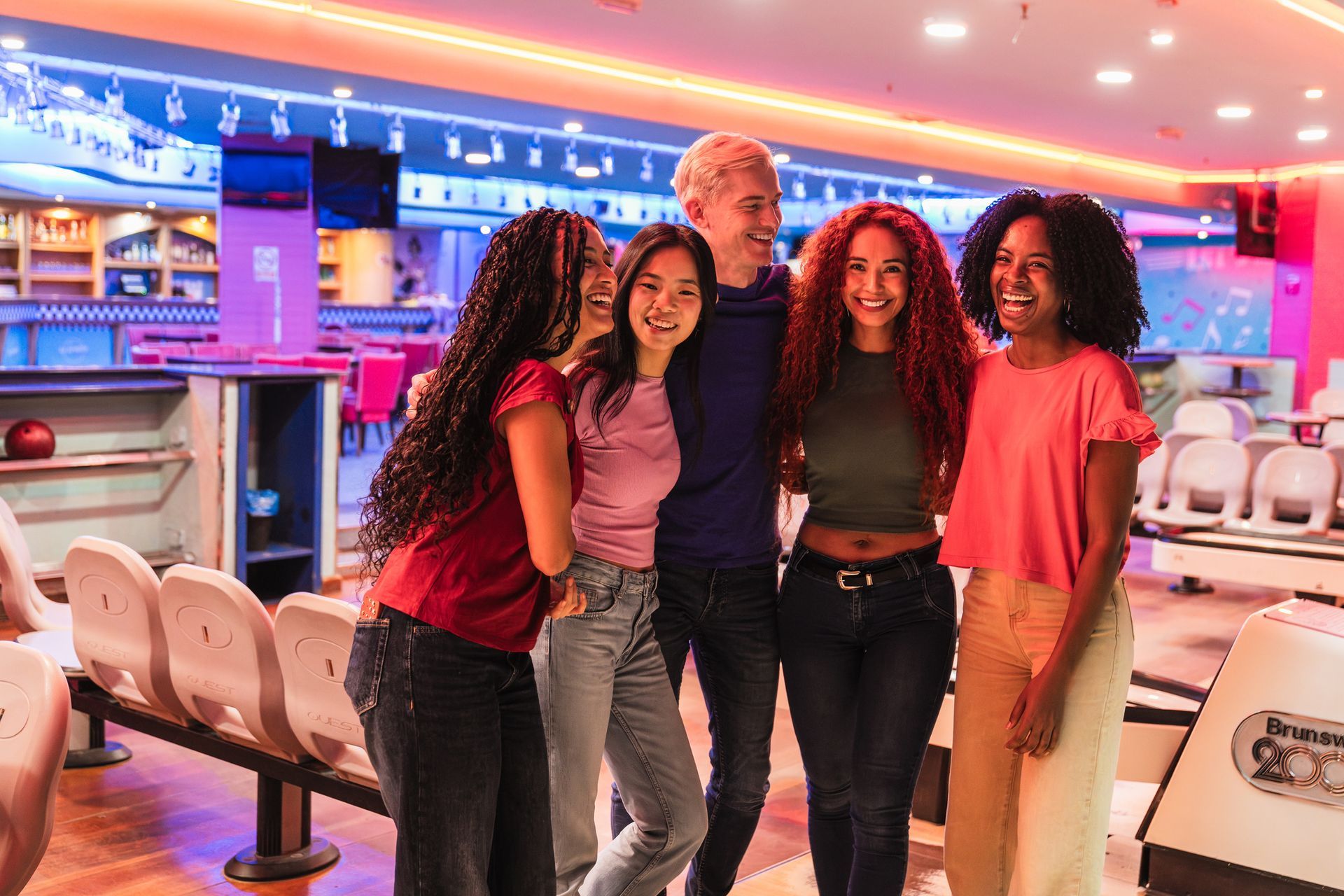 Cinque amici sorridono in una pista da bowling, illuminata da luci rosa e blu.
