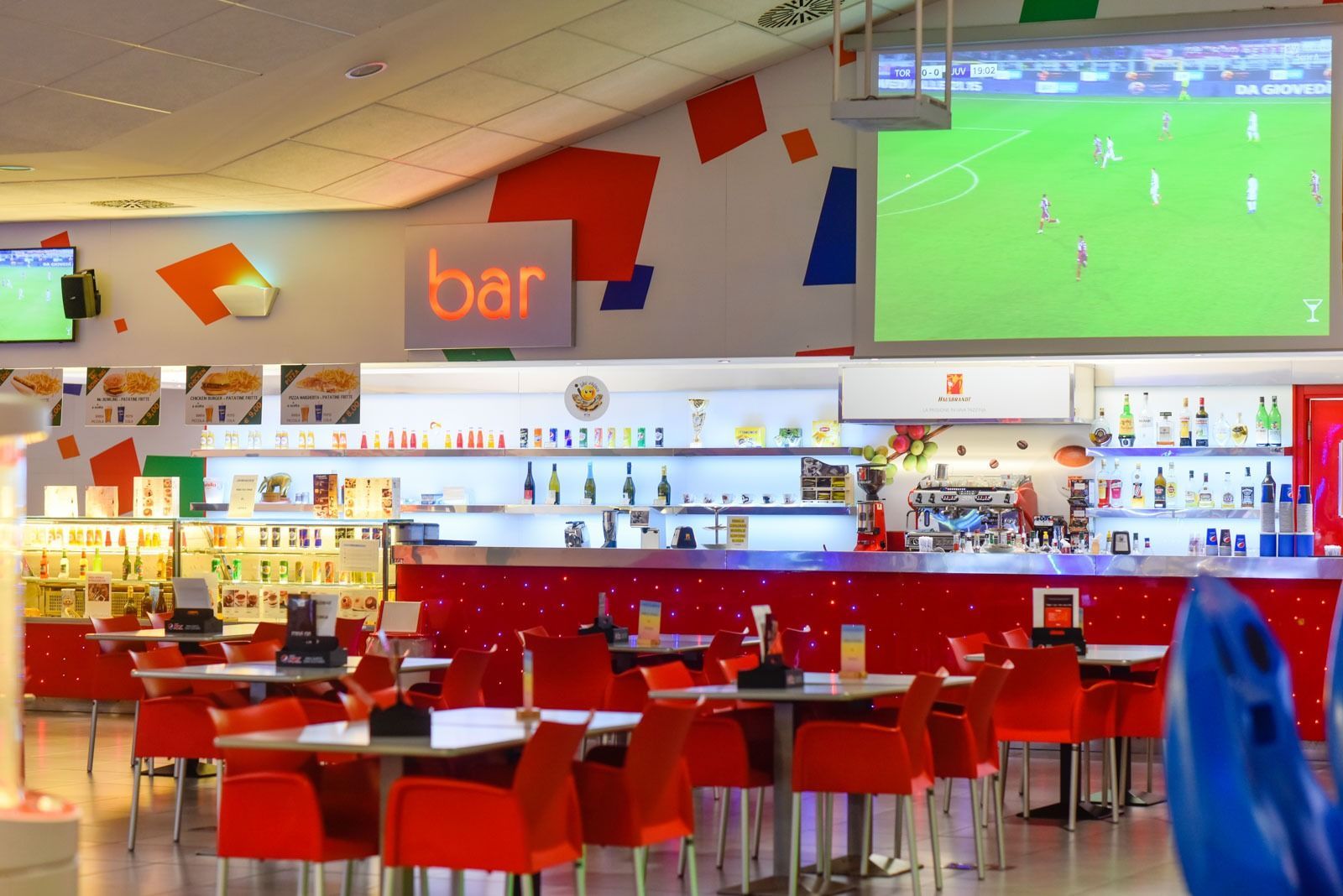 Interno di un bar con tavoli e sedie rossi, un'insegna 