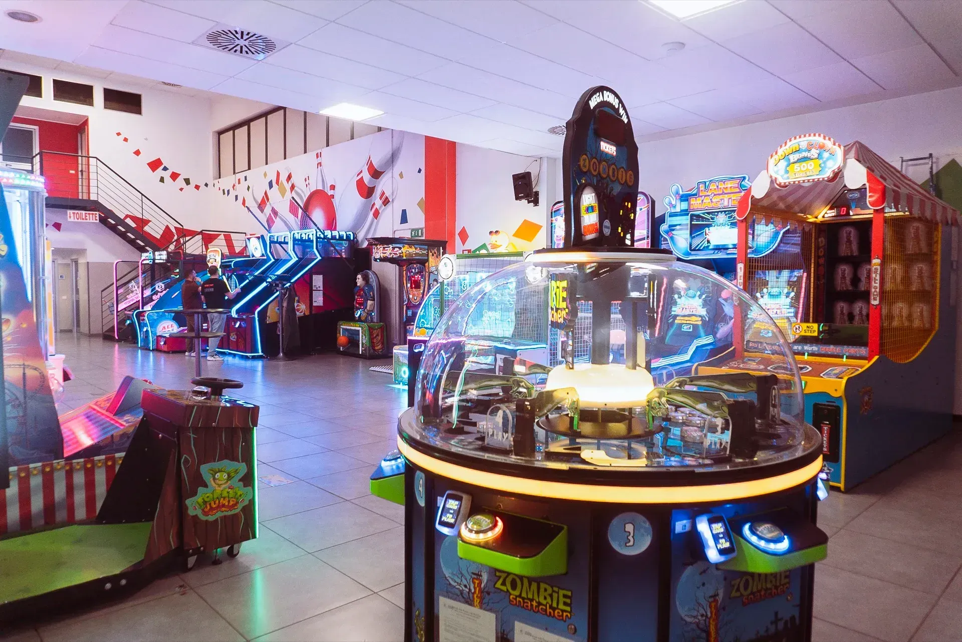 Sala giochi con diverse macchine da gioco ben illuminate. Una scala con dettagli rossi conduce al piano superiore.