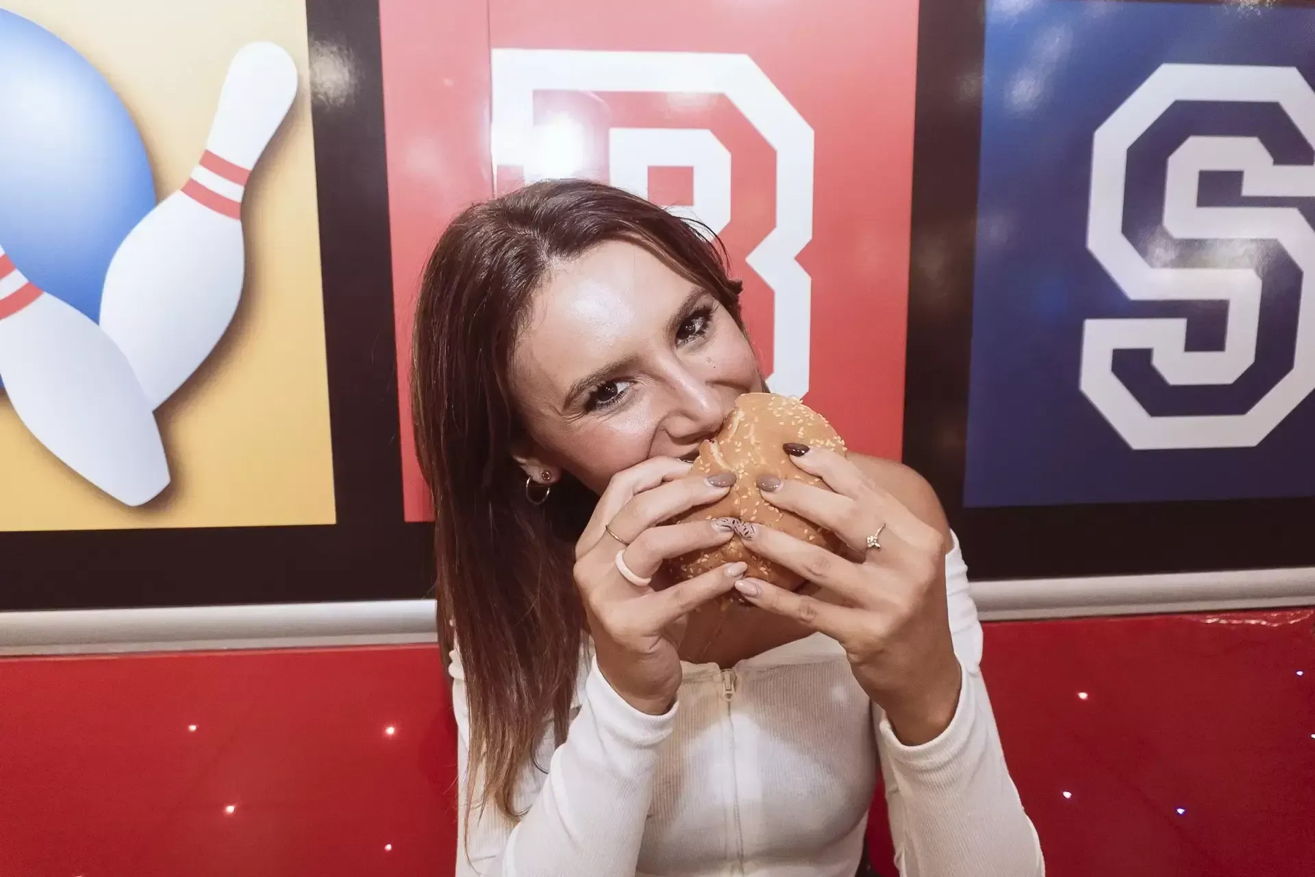 Donna che mangia un hamburger, sorridendo alla telecamera, in un tavolo da pranzo arredato come un bowling.