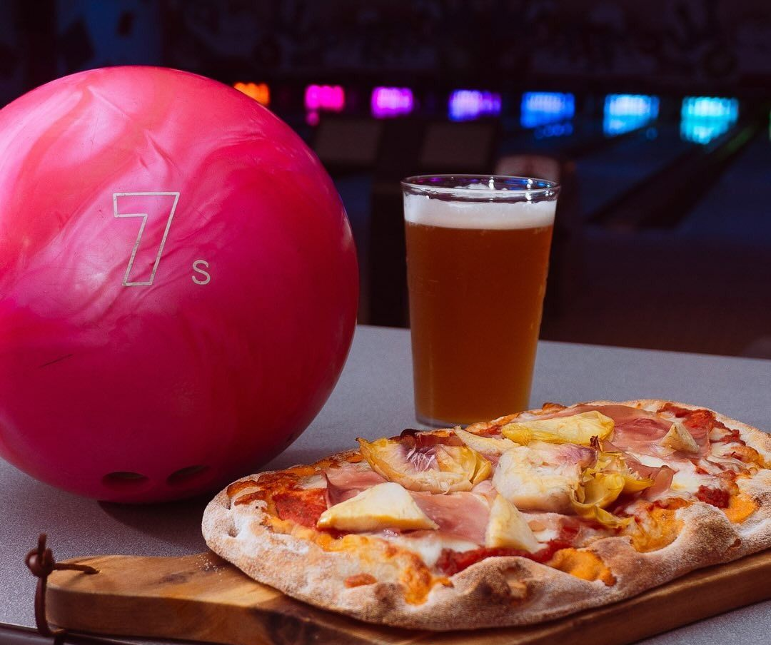 Palla da bowling rosa, bicchiere di birra e pizza su una tavola di legno in una pista da bowling.