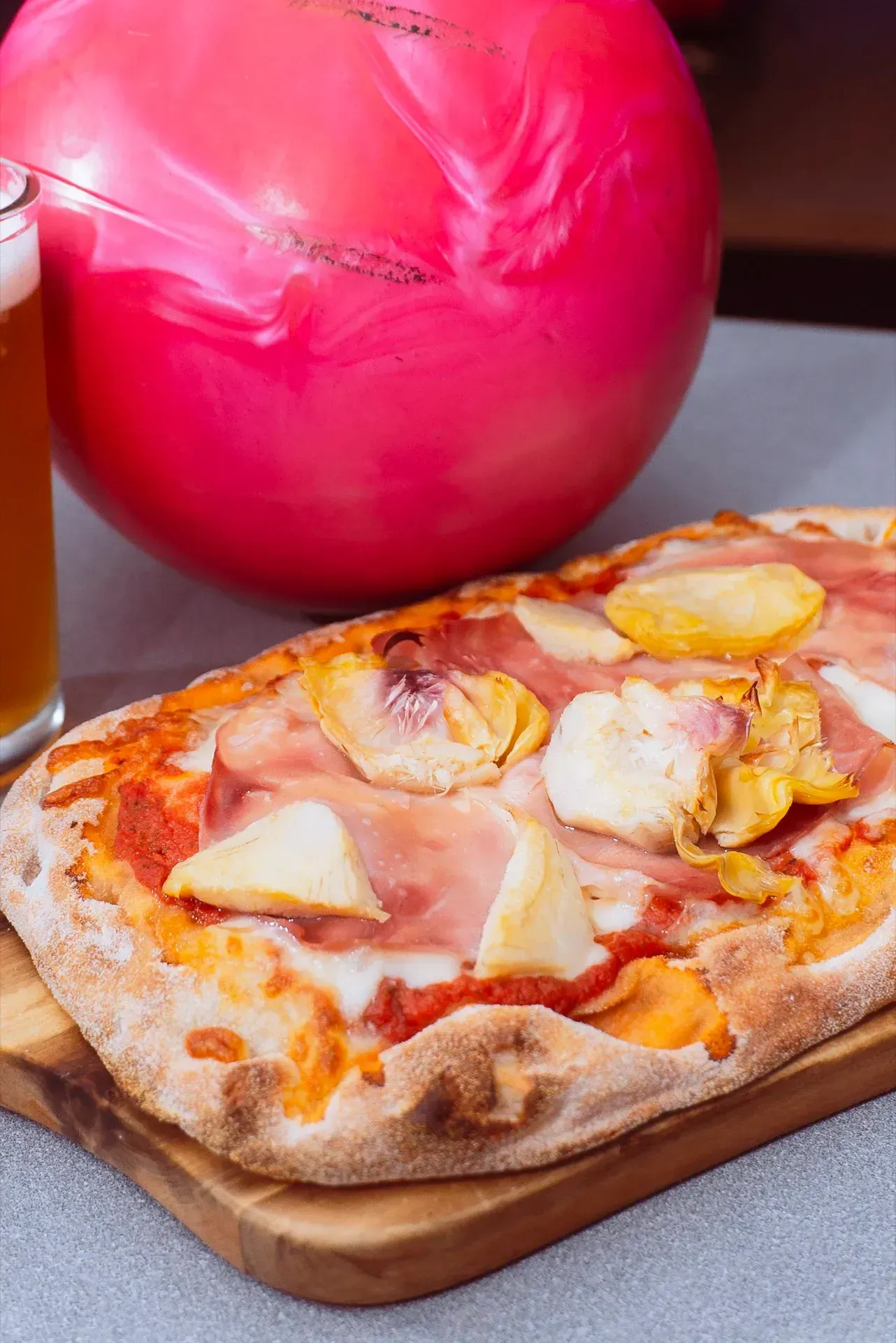 Pizza con prosciutto e frutta, su un tagliere di legno, con una palla da bowling rosa e birra.