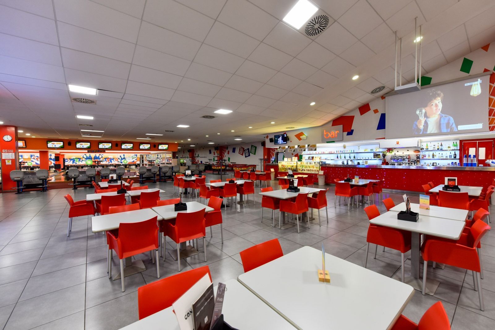 Spazio interno con sedie/tavoli rossi, un bar e TV; probabilmente un ristorante o un'area ricreativa.