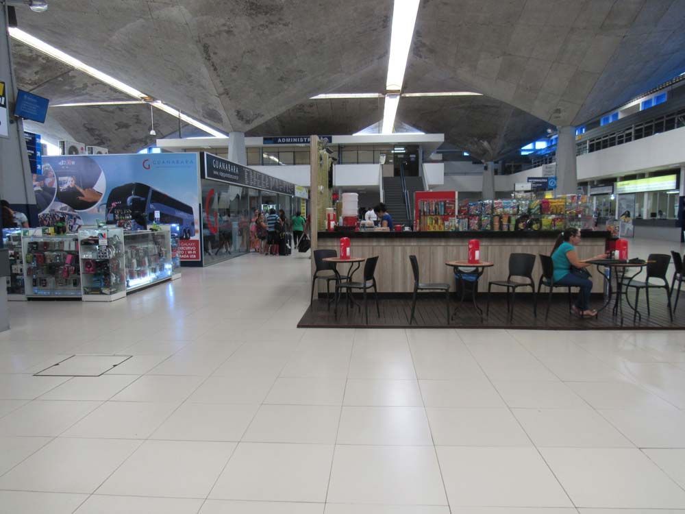 Interior de aeroporto com lojas, mesas de café e teto de concreto. Iluminação intensa.